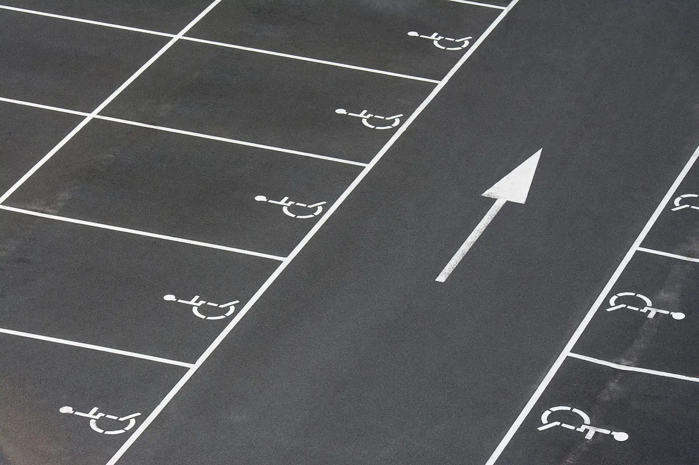 Accessible parking stalls marked out in white paint on dark asphalt, each stencilled with the International Symbol of Access beside a painted directional arrow in the drive aisle