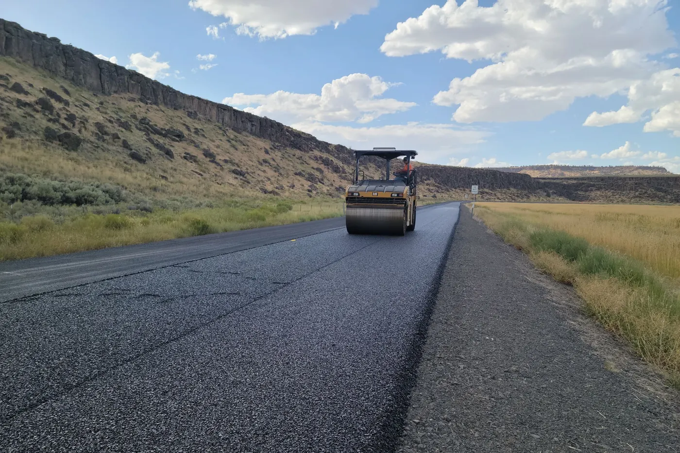 Yellow tandem drum roller compacting a freshly laid black asphalt lane on a two-lane road