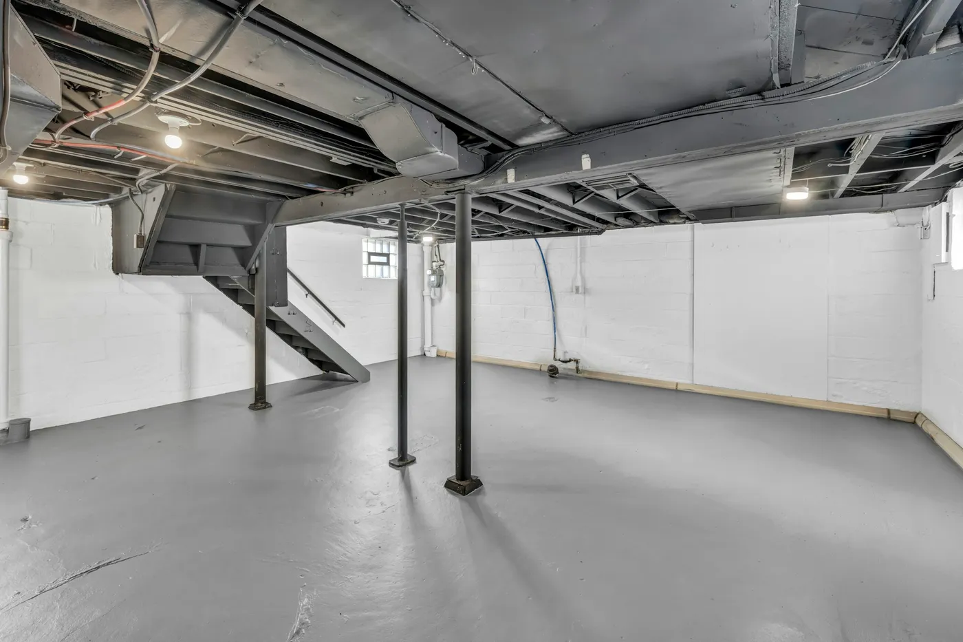 Empty unfinished basement with white-painted concrete block walls, a gray-painted concrete floor, steel support columns and exposed floor joists and ductwork overhead