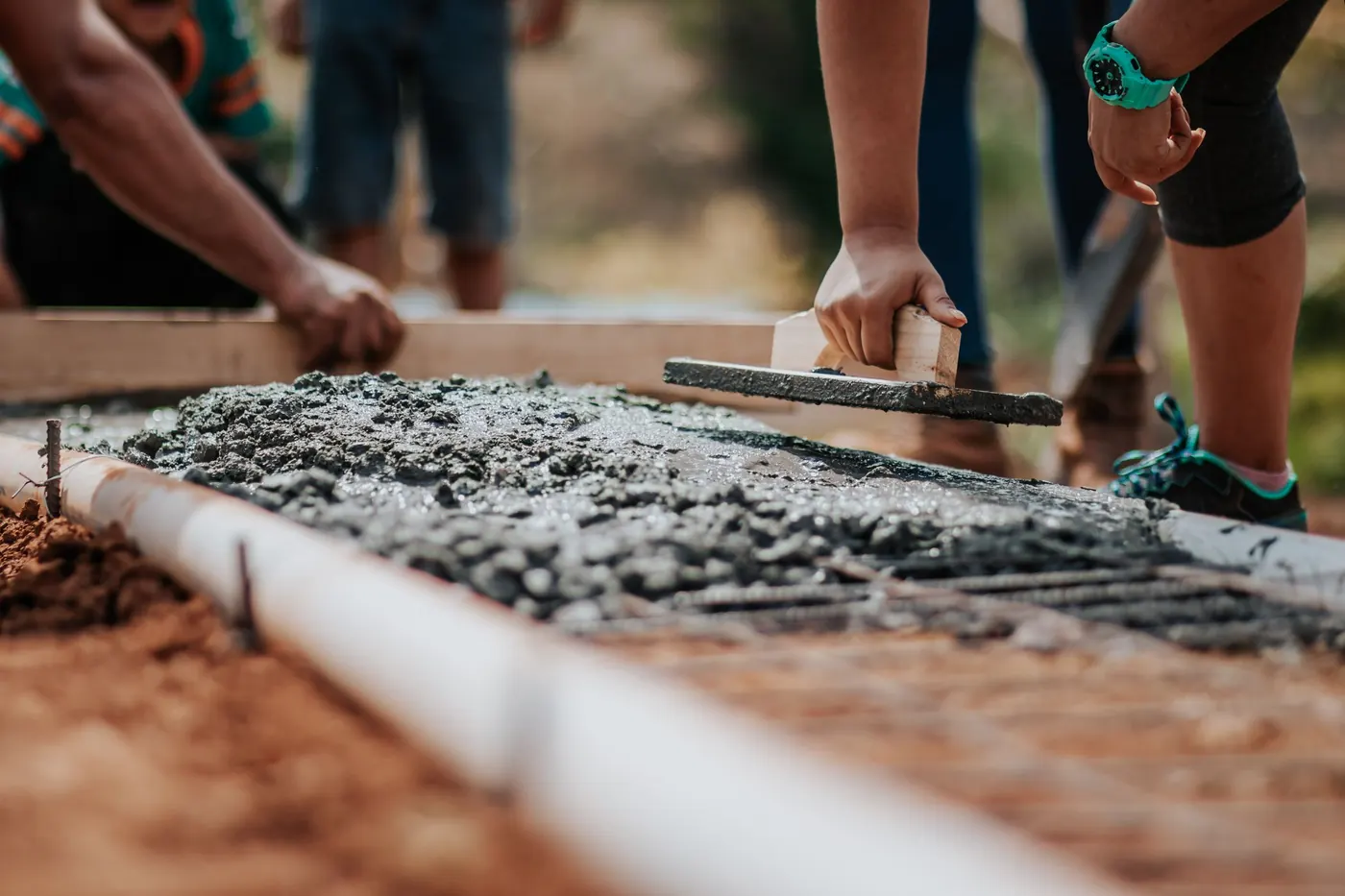 Close-up of a hand holding a steel finishing trowel flat on a strip of freshly placed concrete between timber edge forms, coarse aggregate still showing at the surface