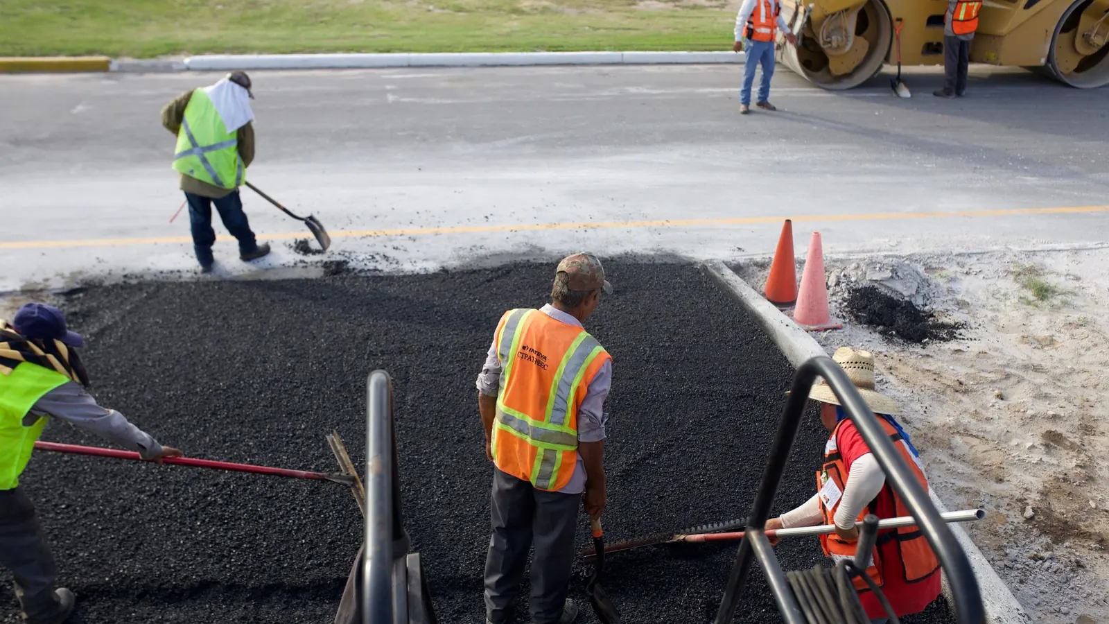 Paving crew in high-visibility vests raking and spreading a fresh mat of black asphalt, with traffic cones and a roller in the background