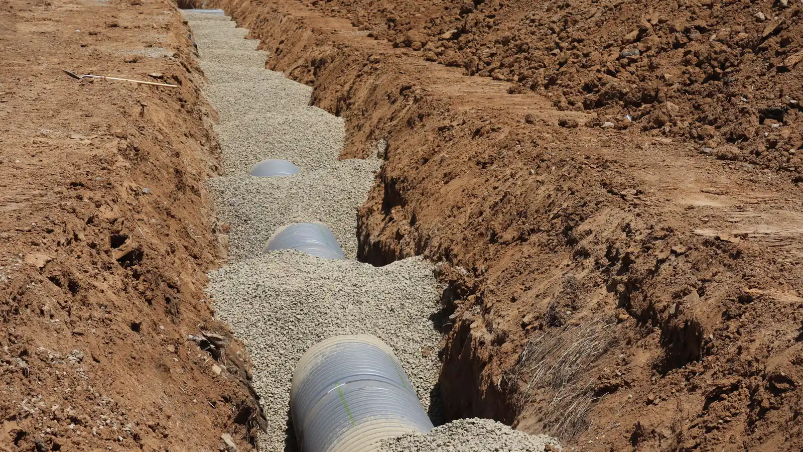 Large corrugated perforated drainage pipe laid along the bottom of an open excavated trench and bedded in clean crushed gravel, with spoil piled along both banks