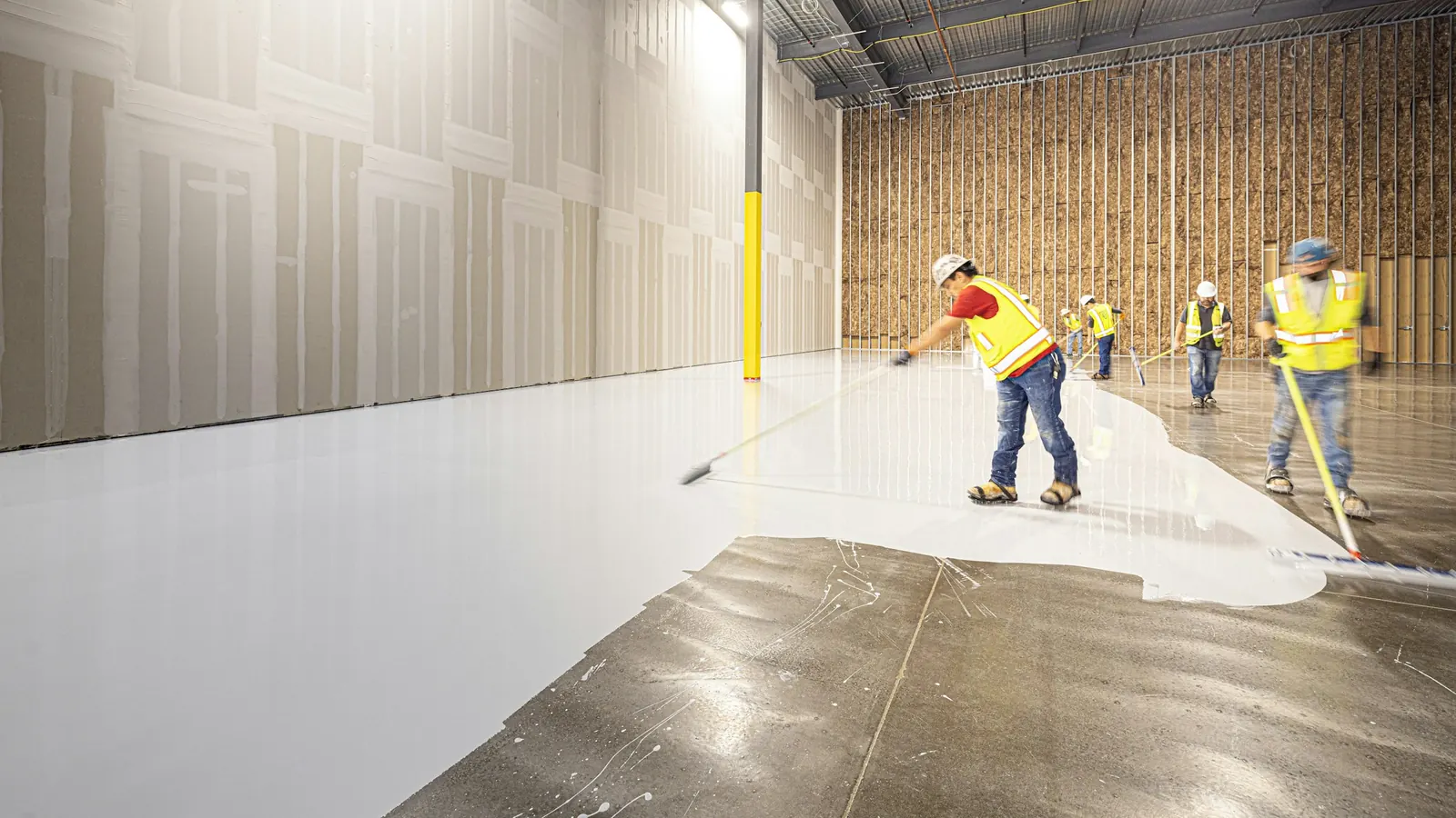 Crew in high-visibility vests spreading a wet light-gray resin floor coating across a bare concrete slab with long-handled squeegees, the coated half glossy and the uncoated half dull