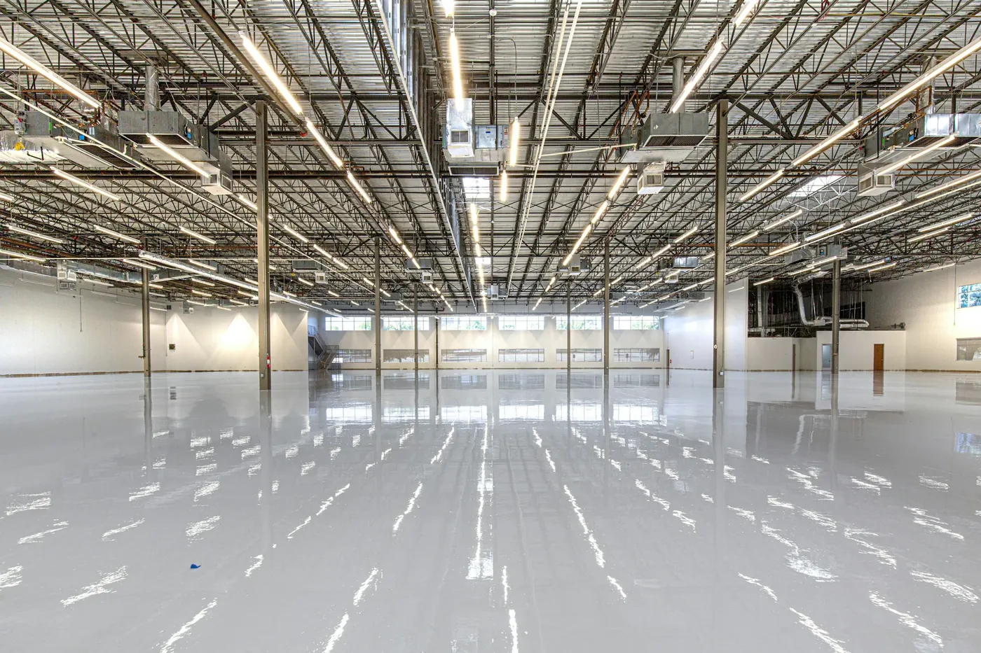 Seamless high-gloss resin floor in a large empty warehouse interior, mirroring long rows of overhead light fixtures beneath an exposed steel joist ceiling