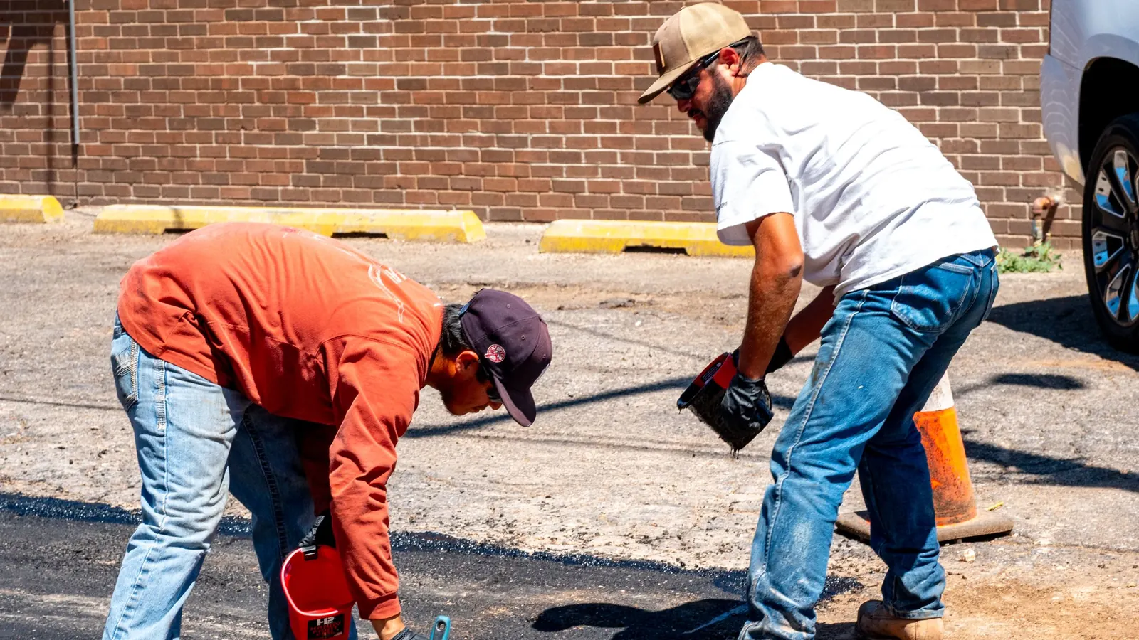 Two workers repairing and sealing a fresh black asphalt patch in a parking lot beside a brick building, with yellow-painted concrete wheel stops along the wall
